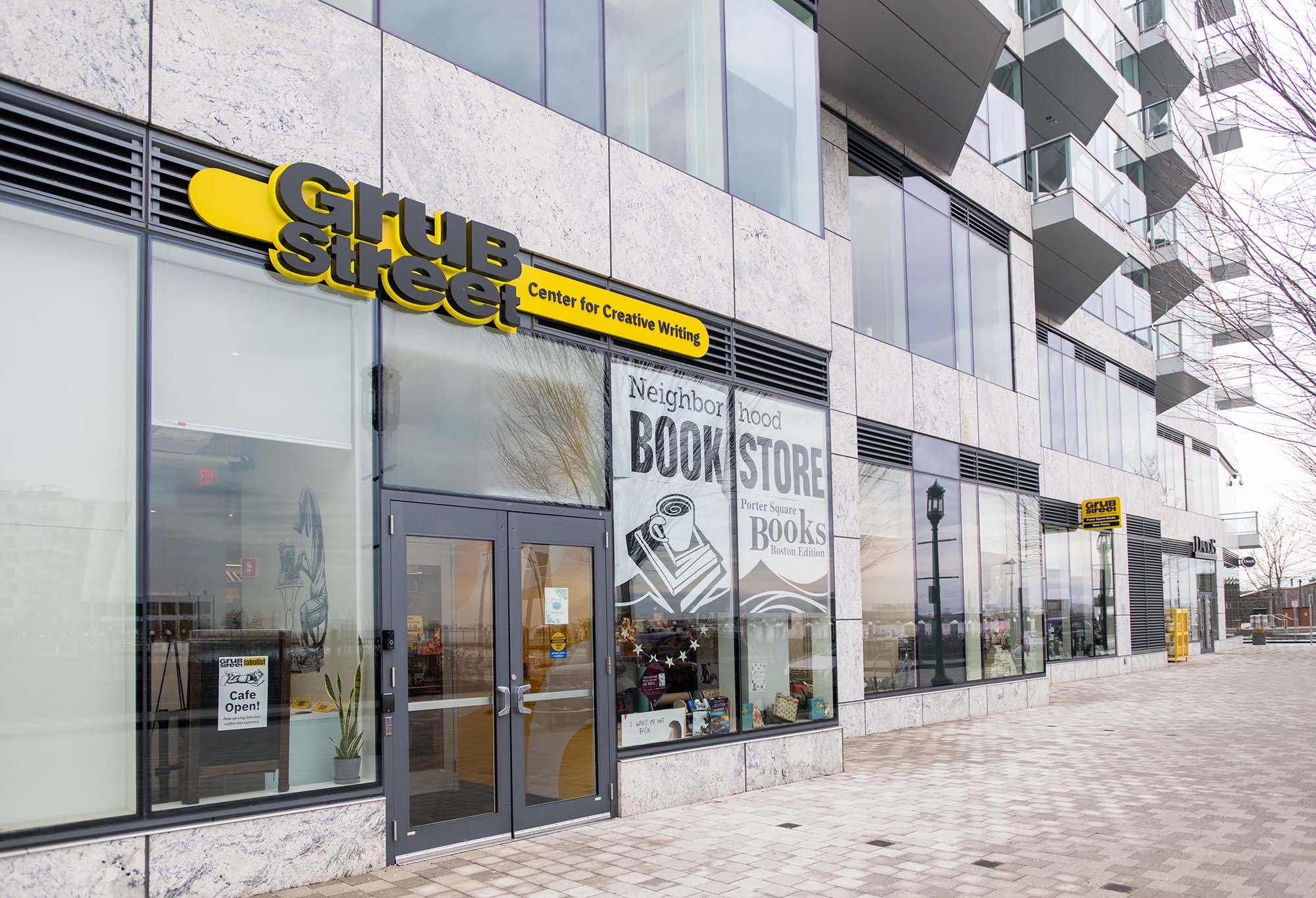 Photo: Exterior shot of a 14-story condominium building on the waterfront. A large yellow sign with black lettering reads "GrubStreet, Center for Creative Writing" above a glass storefront. A large white banner hangs in the store window and reads "Neighborhood Bookstore, Porter Square Books, Boston Edition".