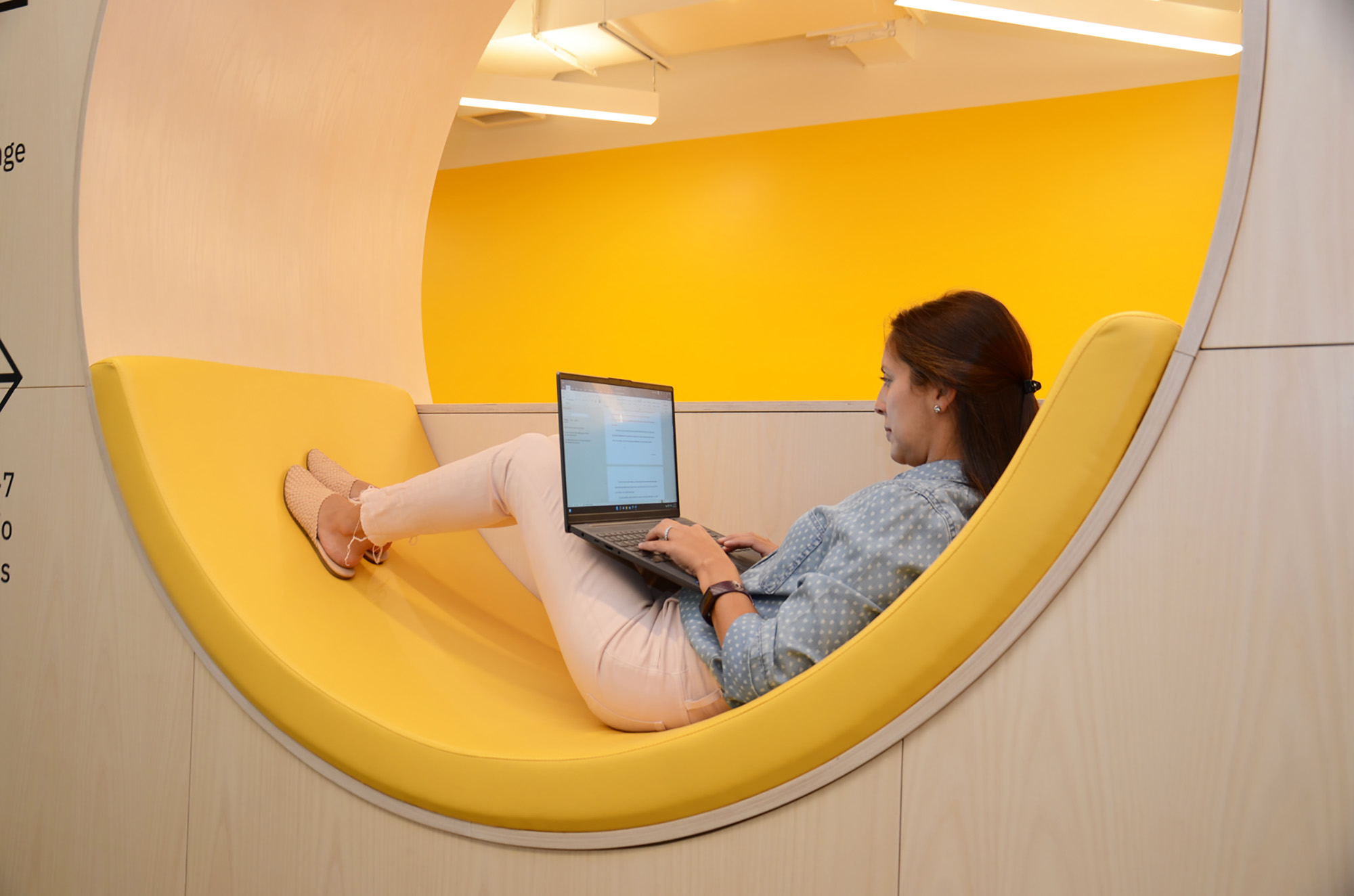 Photo: Shot of one of the many creative space at GrubStreet. A woman lies back in a circular sitting space padded with a yellow seat filler. She works on her laptop in the brightly lit yellow room.