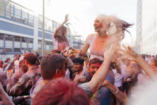 Photo: Vindhya Kotti (MET'23), top, and Varunkumar Gandham Sathish (MET'23), bottom, celebrate Holi at the Cummington Mall on Sunday March 26th. A young woman sits atop the shoulders of a young man as they walk and celebrate in a crowd of people. Everyone is covered in colored powder solutions as colored powder can also be seen throughout the air.