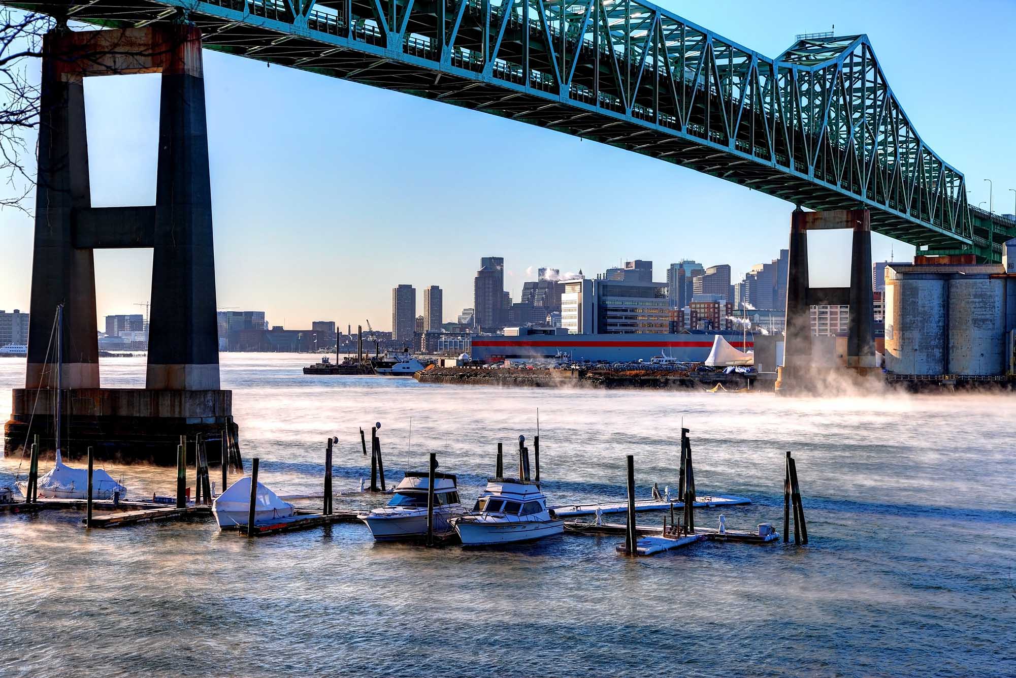 Photo: The Tobin Bridge is shown over the Mystic River in Massachusetts. A large, cantilever truss bridge is shown over a slightly foggy body of water with a small line of boats docked underneath. The city of Boston can be seen in the distant skyline.