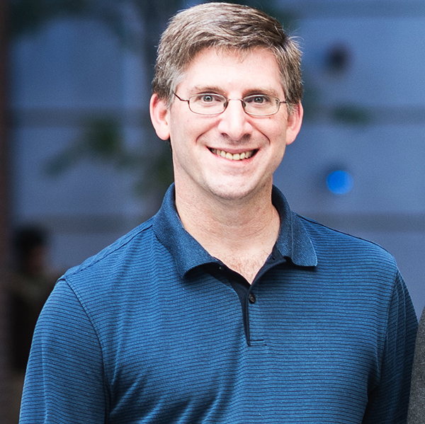 Photo: Portrait of Professor of Environmental Health, Jon Levy. A white man with graying hair and wearing glasses and a blue shortsleeved collared shirt, smiles and poses for the photo. He stands in front of a blue, outdoor backdrop.