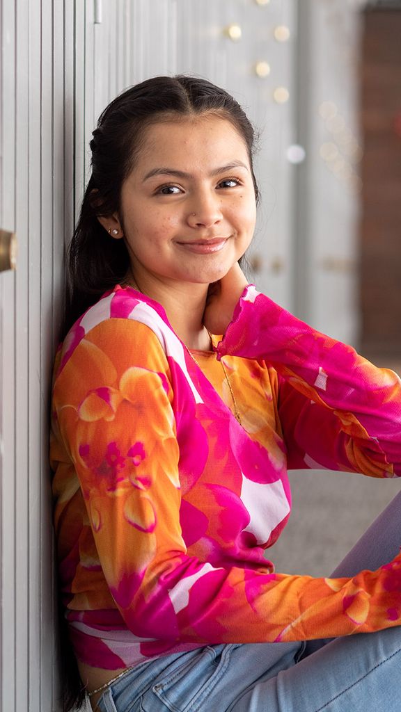 Photo: Kathya Correa Almanza (CAS’25), vice president of Alianza Latina, sits and poses on the floor of a brightly sunlit hallway. A young, Latina woman wearing a pink, orange, and white long-sleeved floral shirt and jeans, poses with one leg bent and arm resting on knee as she smiles for the camera.