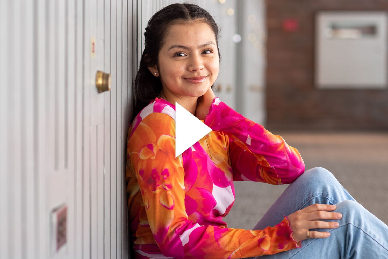 Photo: Kathya Correa Almanza (CAS’25), vice president of Alianza Latina, sits and poses on the floor of a brightly sunlit hallway. A young, Latina woman wearing a pink, orange, and white long-sleeved floral shirt and jeans, poses with one leg bent and arm resting on knee as she smiles for the camera. A white triangle play button is overlaid in the center.