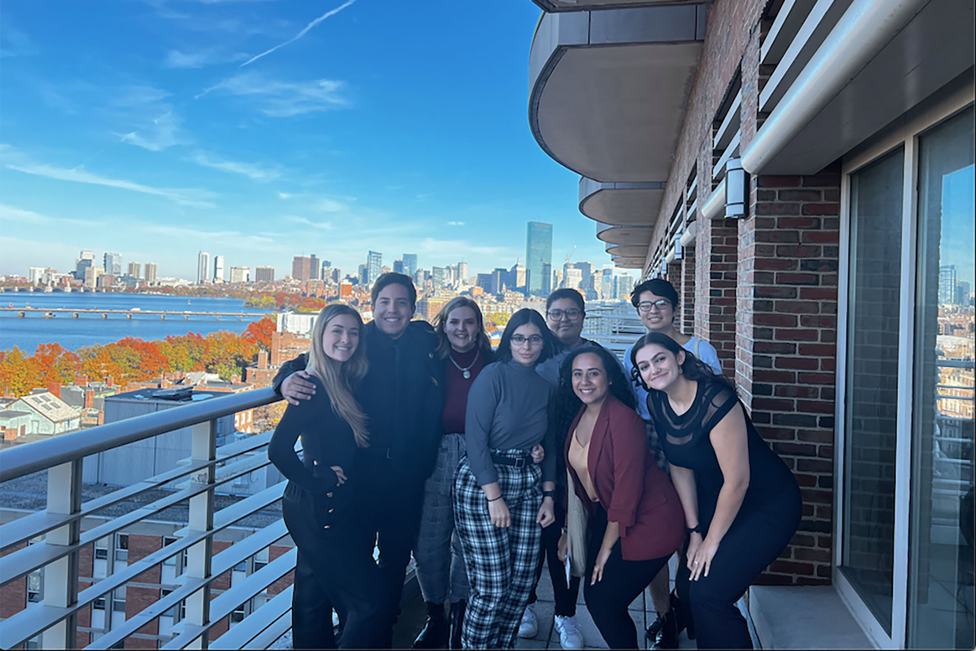 Photo: A group of young adults standing outside a balcony with the Boston skyline in the background. The foliage around them is green, red, and orange, indicating that is is fall outside. The members of the group are all smiling. From left, a blonde woman with straight hair wears all black. Next to her, with his arms around her shoulders, stands a man in all black with a deeper-tone skin and short, dark hair. Next to him is a shorter individual, feminine-presenting with shoulder length hair. She wears a maroon shirt and plaid pants. In front, in the middle of the group, is a woman with short, dark hair, glasses and she is wearing a gray long-sleeve shirt and plaid pants. Next to her are two woman, one with long, curly hair and the other with long, dark straight hair. The curly-haired individual wears a maroon cardigan, a tan shirt, and jeans. The other individual is in all black. Behind the pair is another pair of men, both with short, dark hair and glasses. The entire group smiling.