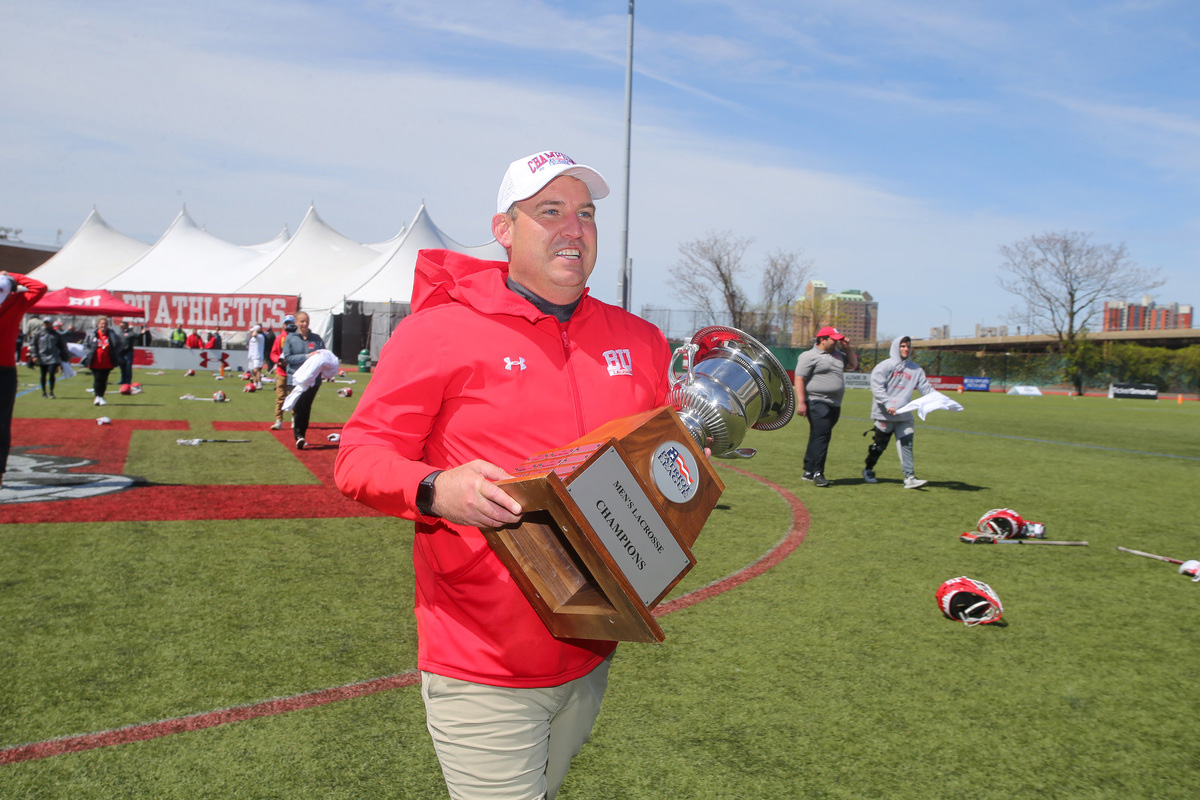 Photo: Terriers head coach Ryan Polley brandishes the 2022 Patriot League trophy, May 8, 2022. An older white man wearing a red BU lacrosse jacket and tan pants smiles as he walks down a green field holding a large silver trophy with a plaque that reads "Patriot League Men's Lacrosse Champions".