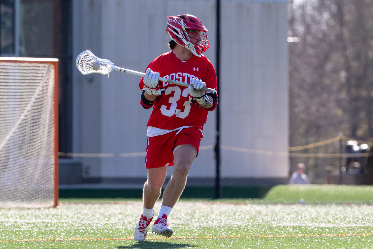 Photo: Captain Matt Hilburn (MET’23), a BU men's lacrosse player in a red lacrosse uniform labeled "Boston 33"and helmet, runs as he holds a lacrosse stick up in the air on the field.