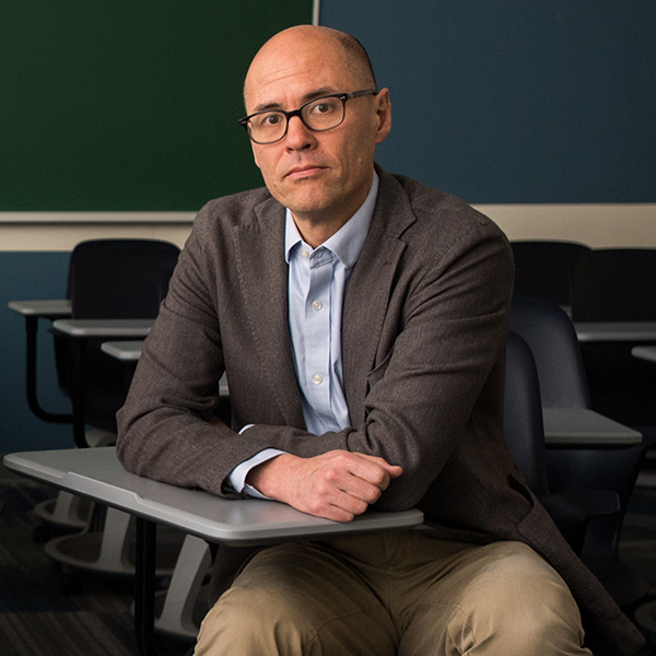 Photo: Professor Maurice Lee sits at a desk with arms crossed and looks tot he camera. A white man with a shaved head and wearing a light blue collared shirt, brown blazer, and tan pants sits and poses in a classroom with empty desks and a green chalkboard in the background.