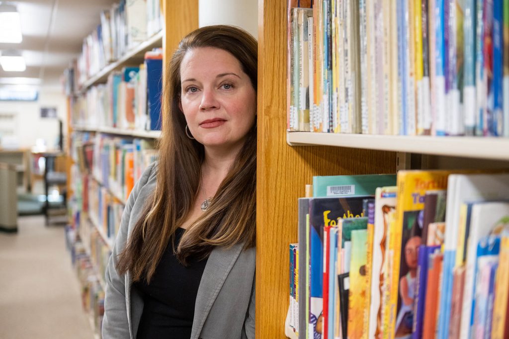 Photo: Portrait of Nancy Nelson. Nelson peeks out from the edge of a wooden bookshelf and poses for the camera. A white woman with long straight brown hair and wearing a black blouse and grey jacket leans against a bookshelf and poses for the camera. Other filled bookshelves can be seen to her left and right.