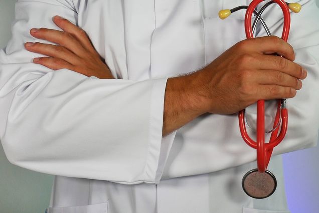 Photo: Stock image of a close-up of arms crossed wearing a doctors white smock. In the right hand, the individual holds a red stethoscope.