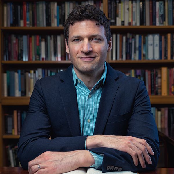 Photo: Joseph Rezek, a white man wearing a teal blue collared shirt and navy blue cardigan, sits with arms crossed on a table in front of him. He sits in front of a bookcase filled with books behind him.