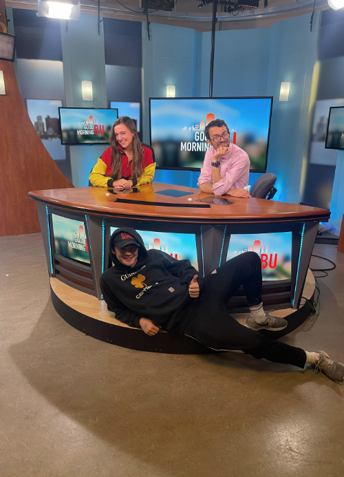 Senior, Kelsey Martirano (COM'23), pictured in the BUTV10 news room, accompanied by two other students. One student is smiling to the right of her in a chair and the other lying below the news desk, giving a thumbs up. 

Picture credit: Kelsey Martirano (COM'23)