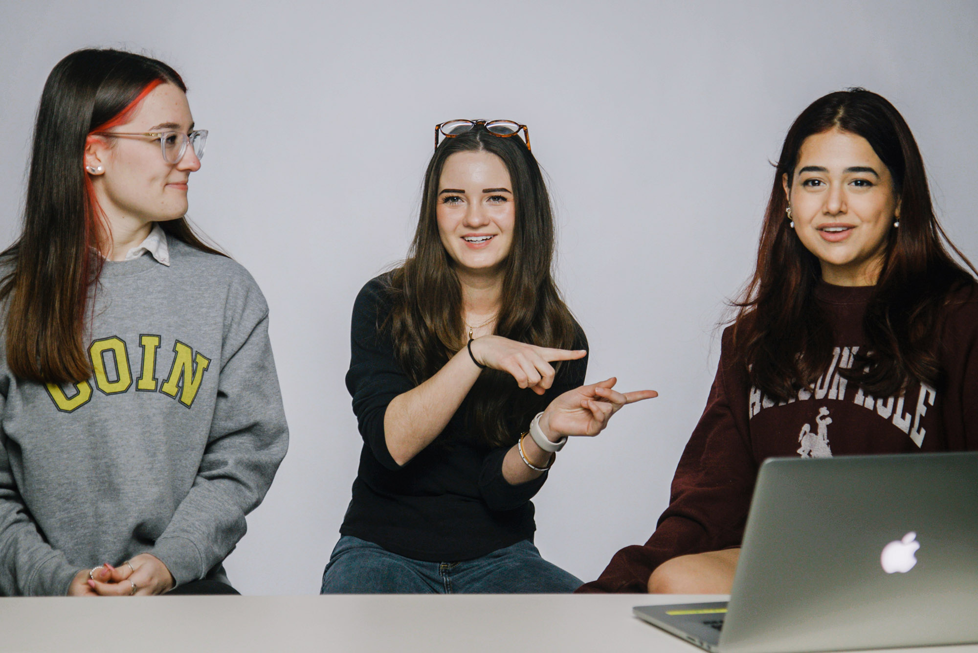 Screenshot of three young white women sitting at a table in front of a light grey backdrop. The woman on the left wears glasses and a grey sweatshirt and looks at the woman in the center. The woman in the center wears a black shirt and jeans with glasses resting on top of her head. She pint to her right towards the woman on the right. The woman on the right wears a black sweatshirt and looks straight ahead to the camera and is caught mid-speaking. An Apple laptop lays open in front of her.