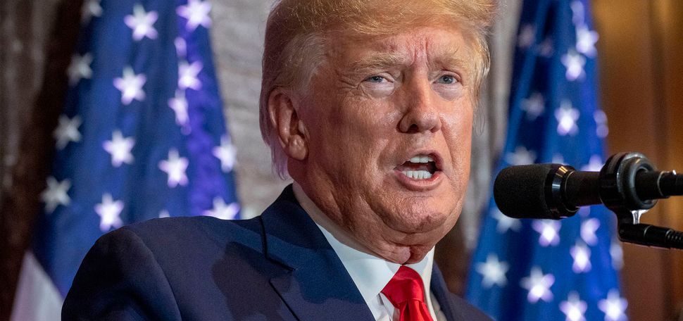 Photo: Candid photo of Donald Trump speaking. An older white man with an orange-tinged tan and wearing a navy suit and red tie speaks into a microphone. A US flag is seen behind him.