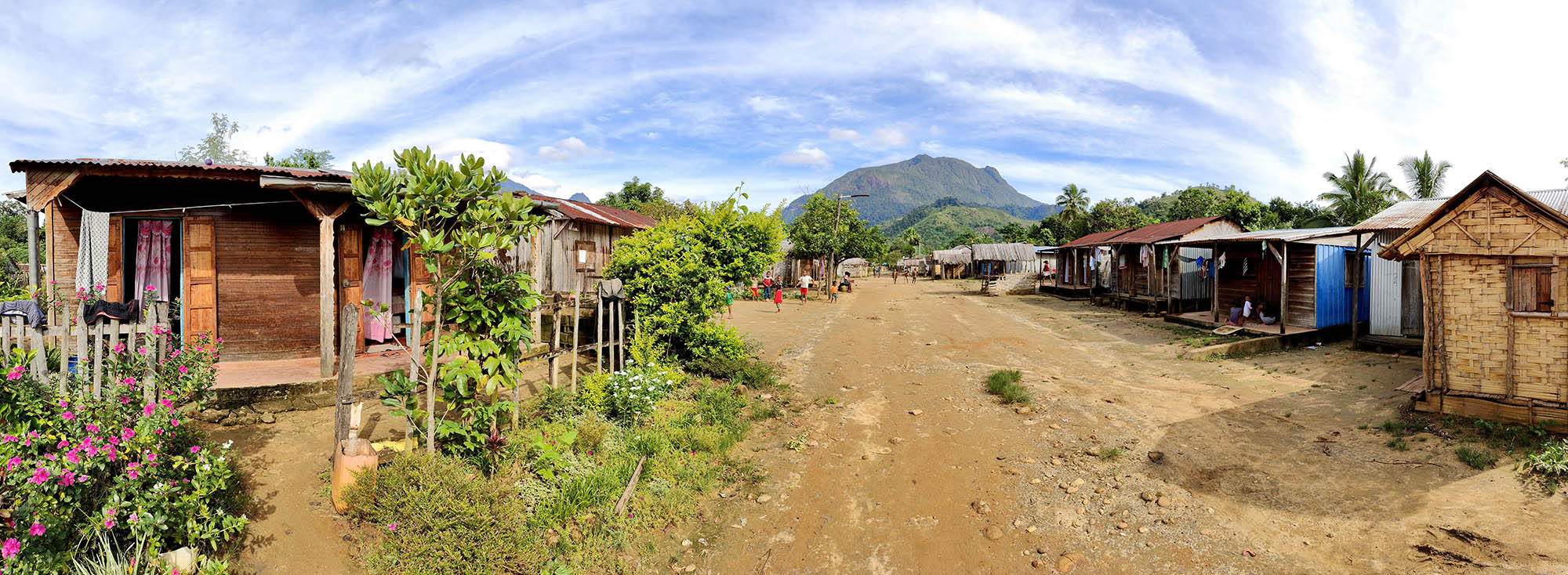 Photo: A rural village farm in Africa. A dirt road with greenery and small, wooden houses are shown on either side. Children can be seen playing in the distance as chickens wander around. A large green mountain can be seen in the distant background.