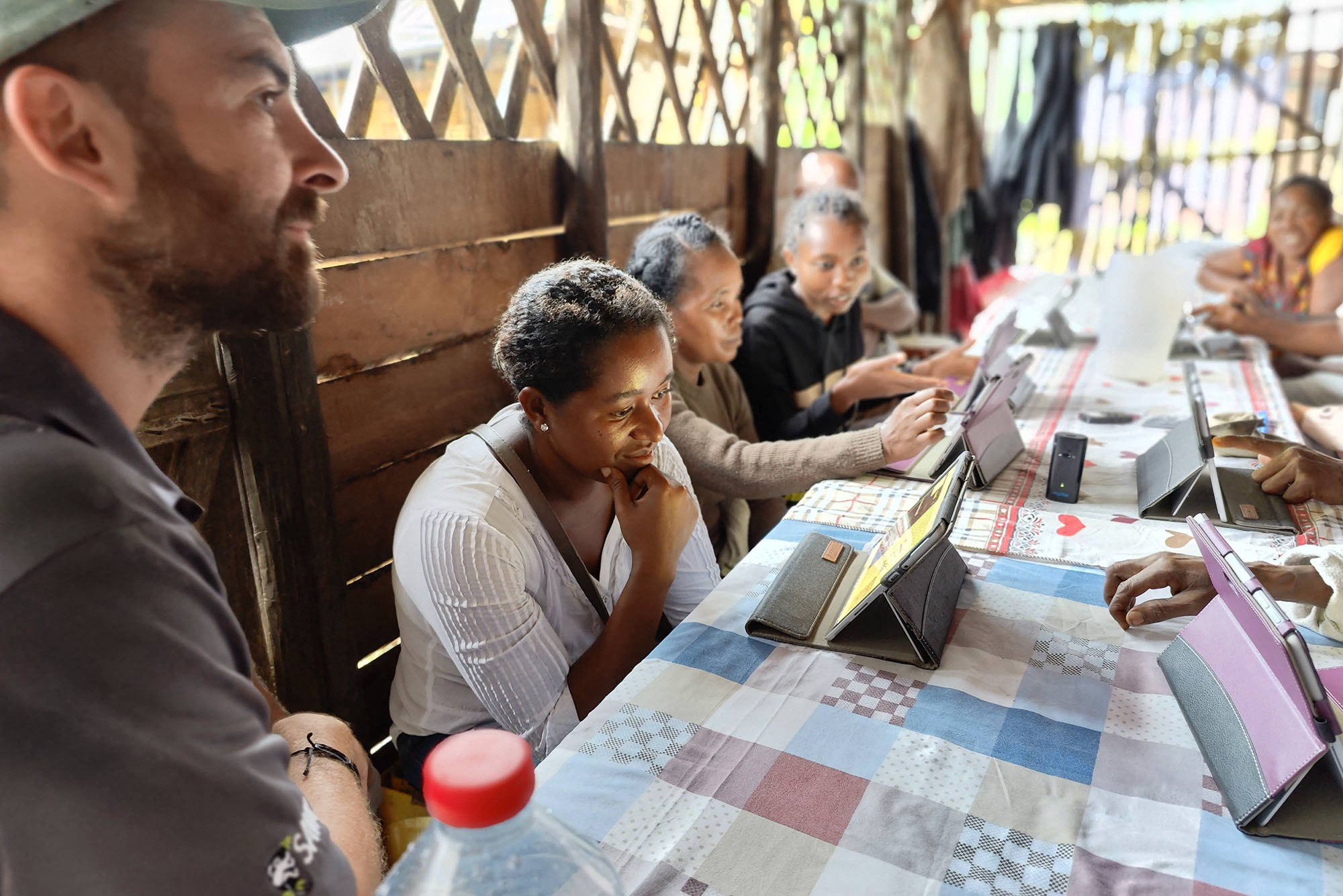 Photo: To the far left, Andrew Reid Bell, a white man wearing a green cap, looks on as a small group of dark-skinned Black women and men look to tablets on a multi-colored, checkered long table. They are in a small wooden dwelling with an open doorway and cross-patterned open windows letting light in.