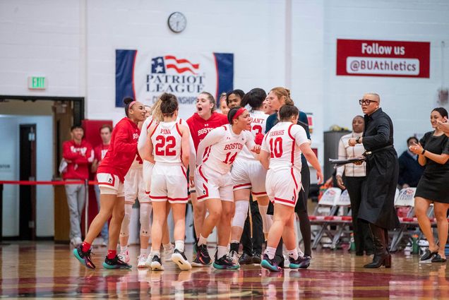 Photo: A group huddle of BU women's basketball. They all wear their white uniforms with red lettering. They are all celebrating.