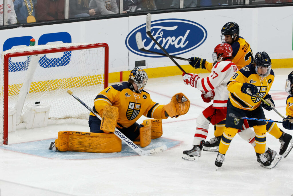 Photo: The Merrimack goalie attempts to defend BU hockey player as he tries to make a goal. Merrimack uniform is gold and black details while BU is white with red detailing.