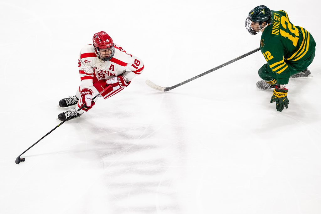 Photo: A BU Hockey player battles aganist another player on the ice. The BU player is wearing their uniform with white background and red lettering. The other player wears a green uniform and yellow lettering.