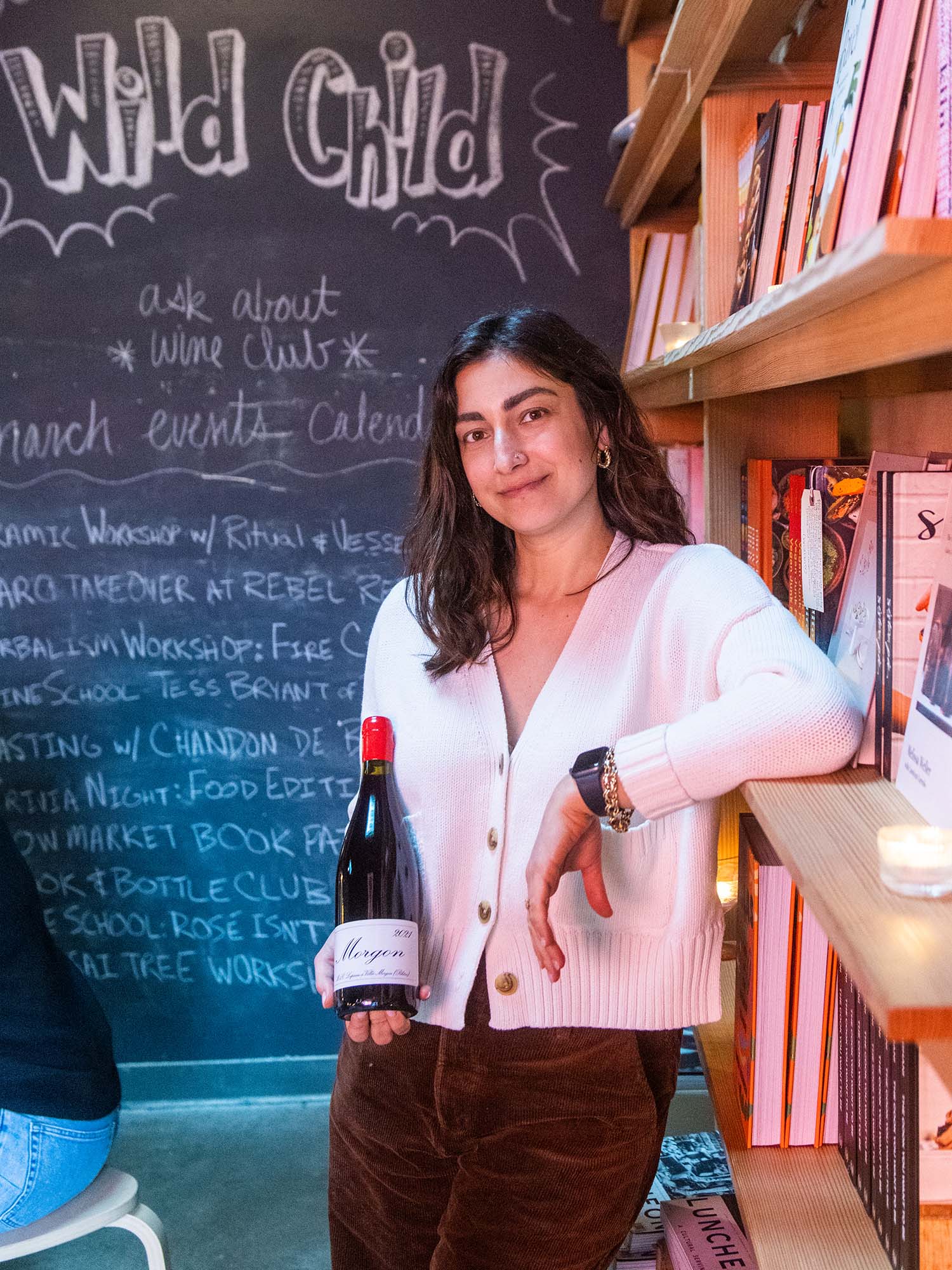 Photo: Friedland holds a bottle of red wine, relaxing on the bookshelf with a chalkboard wall behind her. She wears a white button up shirt, black pants, and has short brown hair. The largest letters behind her read "Wild Card".