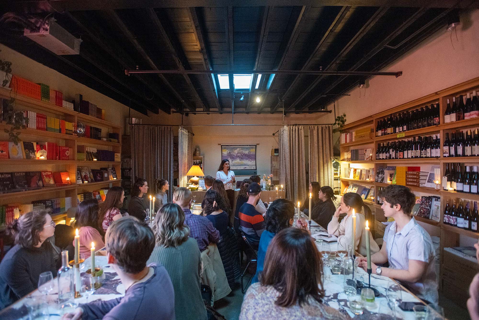 Photo: A crowd of people sit and listen to Friedland in the background of the photo. There are two, long horizontal tables with a tablecloth and candlesticks which are lit. The crowd sits in a small room.