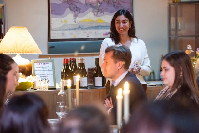 Photo: Friedland, a woman with shoulder-length black hair and a white button up top on, stands over the crowd at her wine bar, Rebel Rebel. She is smiling. The table is set with a tablecloth and candlesticks that are lit. A white man sits in front of where she stands and looks across the table.