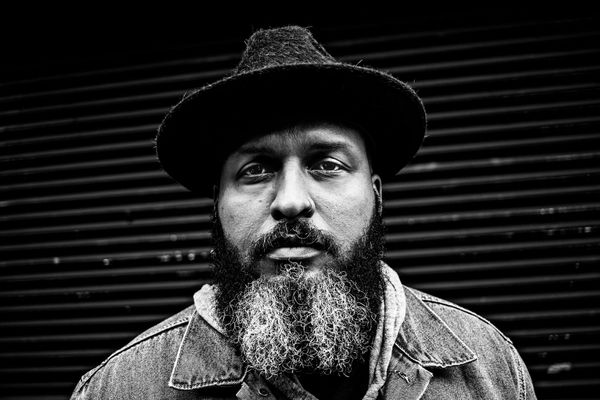 Photo: A black and white photo of a Black man with a long, peppered beard. He has a nose piercing in his left nostril. He was a collard shirt, a sweater overtop, and a flat-brimmed hat.