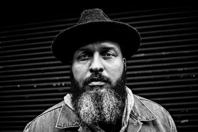 Photo: A black and white photo of a Black man with a long, peppered beard. He has a nose piercing in his left nostril. He was a collard shirt, a sweater overtop, and a flat-brimmed hat.