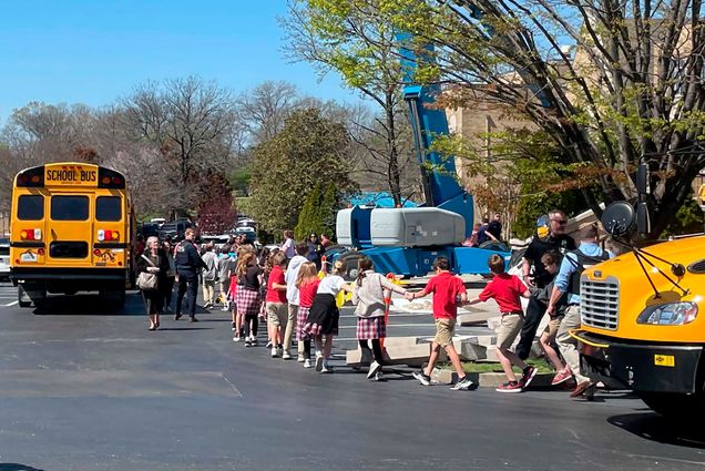 Photo: A line of children hold hands as they are ushered out of their school after a school-shooter threat. They were variations of their school uniform and are walking towards a school bus.