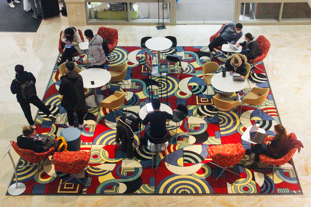 Overhead photo of students at Questrom School on Wednesday, January 26, 2022. The students sit a circle tables in small groups in bright red chairs. The tables and chairs are on a patterned rug with a series of circles and other geometric shapes.