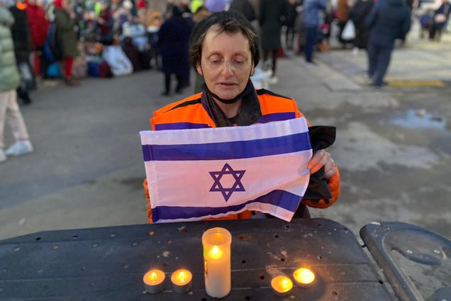 Photo of Jill Shames (COM’82 SSW’83), lighting Shabbat candles and holding a small Israeli flag, after helping Ukrainian refugees from the Russian invasion as they enter neighboring Moldova. She looks solemn, wears a hat, a bright orange vest and stands at a black table.