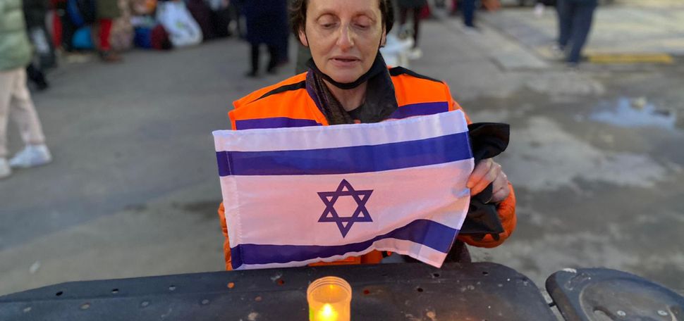 Photo of Jill Shames (COM’82 SSW’83), lighting Shabbat candles and holding a small Israeli flag, after helping Ukrainian refugees from the Russian invasion as they enter neighboring Moldova. She looks solemn, wears a hat, a bright orange vest and stands at a black table.