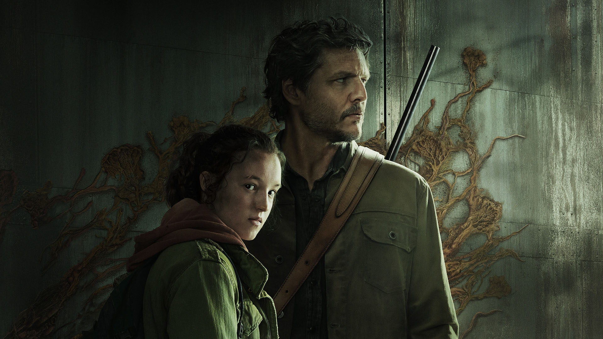 Image: Promotional image for the HBO drama series "The Last of Us". Image shows Joel, played by Pedro Pascal, and Ellie, played by Bella Ramsey, looking stoically into the distance. Photo is tinged in a sickly green and both characters are dressed in grungy layers of clothes.