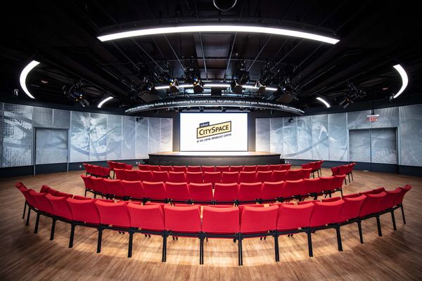 Photo: A wide shot of a production center with rows of red chairs facing a huge stage. They sit on a wooden floor. The screen in front of them is title WBUR CitySpace. The room is dimly lit.