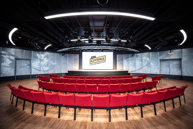 Photo: A wide shot of a production center with rows of red chairs facing a huge stage. They sit on a wooden floor. The screen in front of them is title WBUR CitySpace. The room is dimly lit.