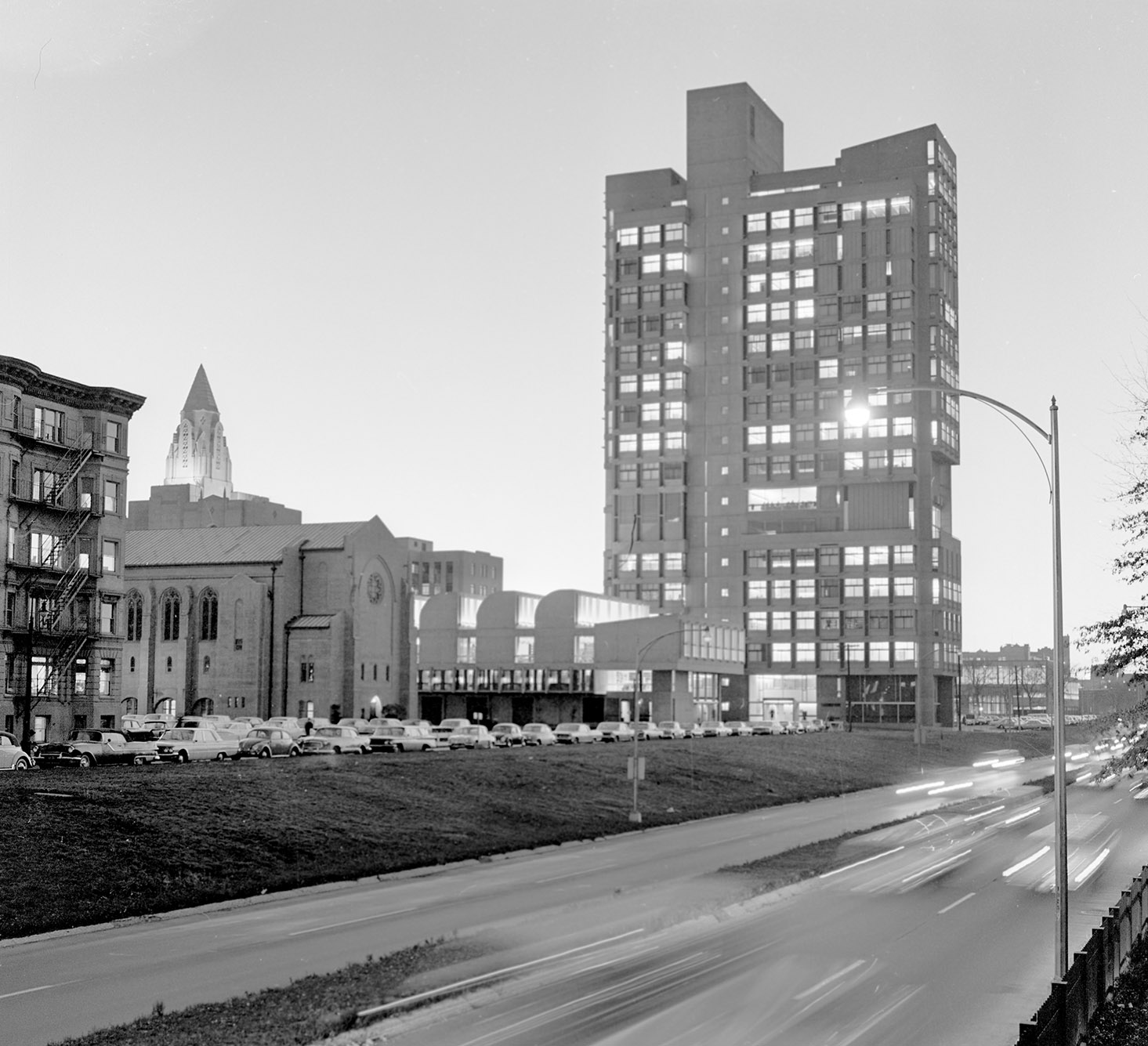 Photo: Black and white photo shows sunset traffic passing Boston University on Storrow Drive in 1964. A large lit building is shown next to a whizzing highway as old school cars are parked on the lawn nearby.