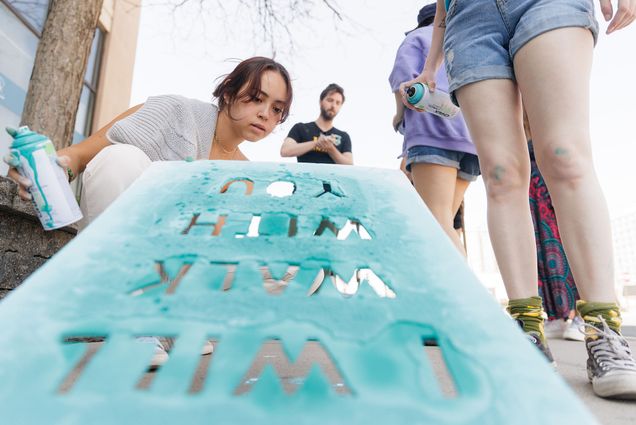 Photo: BU SARP Ambassador Sarah Annis (QST'25) participated in the "I Will Walk With You," public art event in which students spray painted footprints on the sidewalk to symbolize community support for victims of sexual assault on Thursday morning. A young white woman wearing a light grey sweater vest holds up a light blue, stained stencil that reads "I will walk with you". Next to her the legs of another student are shown as well as their hand holing a can of light blue spray paint. A few other students can be seen in the blurred background behind them as they work to stencil the sidewalk.