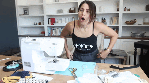 This is a GIF of a woman in front of a sewing machine and work station, expressing excitement. She is a smiling and punches the air with in celebration.