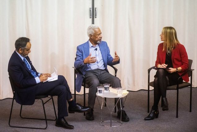 Photo: Abdulrazak Gurnah , Professor Muhammad Zaman (left) and Professor Carrie Preston sit at the front of Morse Auditorium on April 24, 2023. (from left) a tan man wearing a light blue collared shirt and navy blue suit looks and writes on sheets of paper, a Black man with white hair and a gray trimmed beard wearing a light blue collared shirt, blue blazer, and dark grey pants is turned to his right as he talks and gesticulates to a white woman wearing a red blazer, black dress, and black tights. They all sit in black chairs in front of a white background. A small table is at the center of them holding small plastic cups of water.