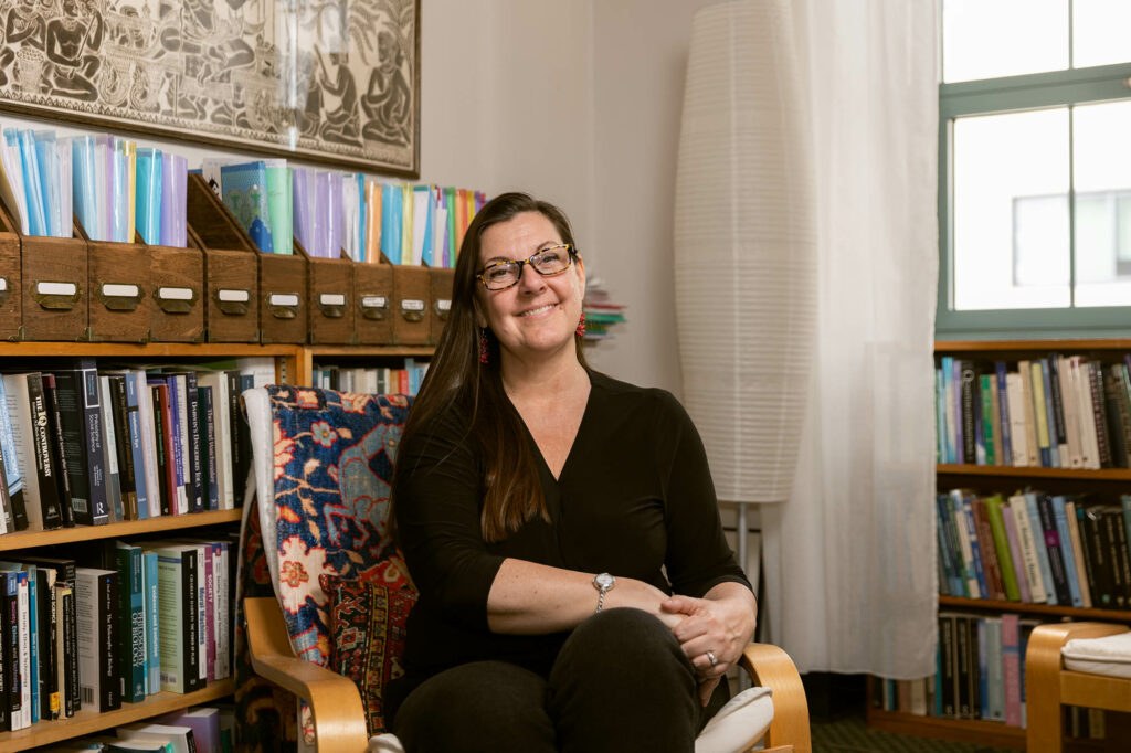Photo: Dr. Bokulich, a white woman with long, dark brown hair and wearing a long black dress with long sleeves, sits and poses in her cozy office. Bokulich stands in an armchair with a floral blanket draped on the seat. She seats in front of a small, filled bookcase.