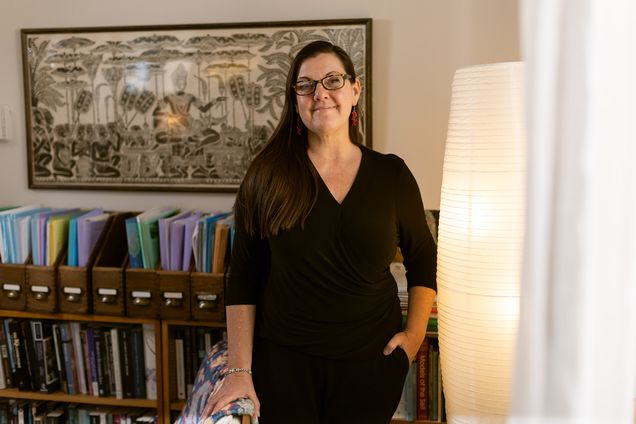 Photo: Dr. Bokulich, a white woman with long, dark brown hair and wearing a long black dress with long sleeves, poses in her cozy office. A tall warm-light lamp lights the room as Bokulich stands and poses next to a floral armchair and in front of a small, filled bookcase.