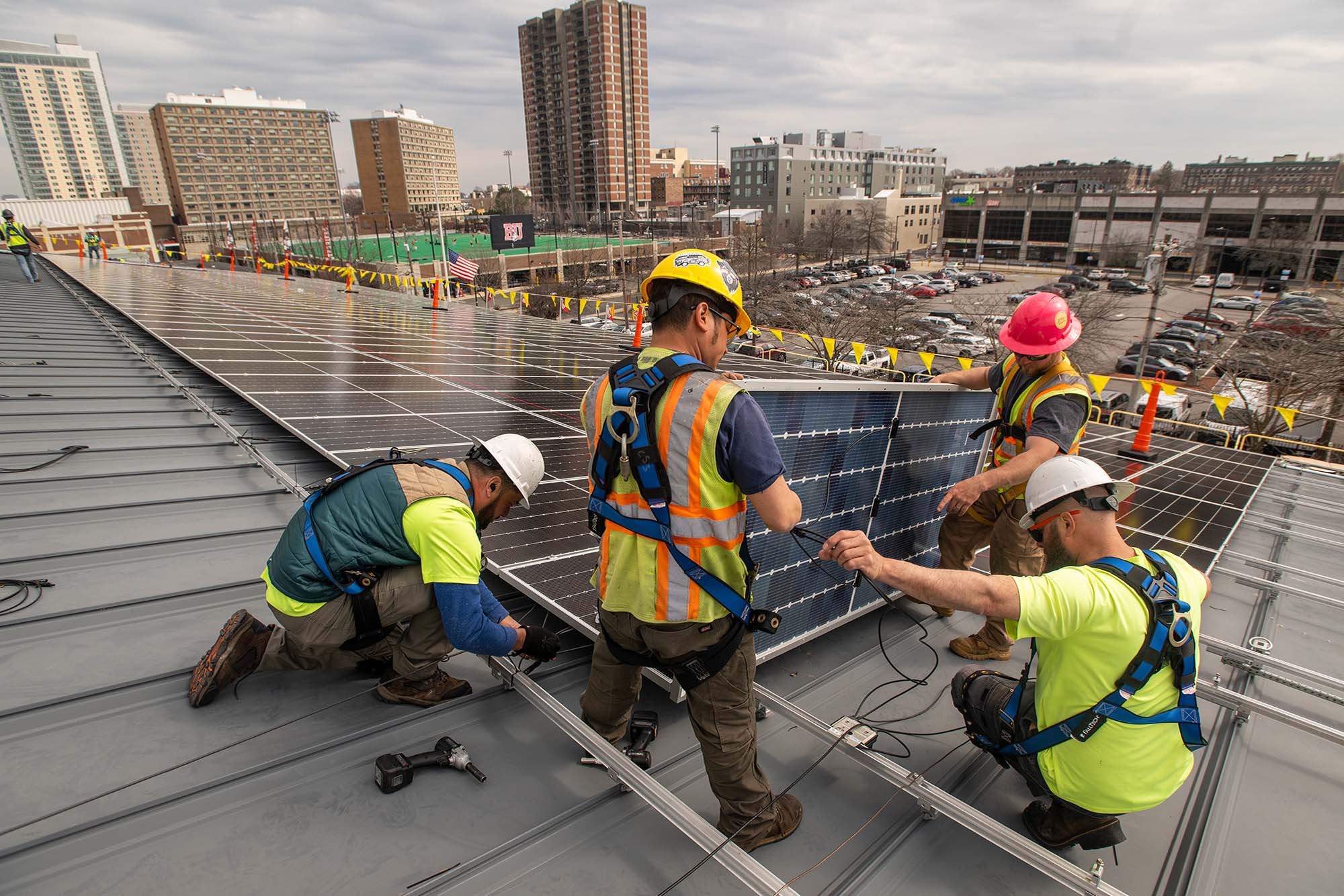 Photo: A team from Solect Energy instals what will ultimately be 1500 solar panels on the roof of the Track and Tennis Center April 6. A group of men wearing hard hats, neon vests, and construction gear work to place a large solar panel on the roof of a large building. In the distance the New Balance field, BU's West Campus, and various brick buildings can be seen.