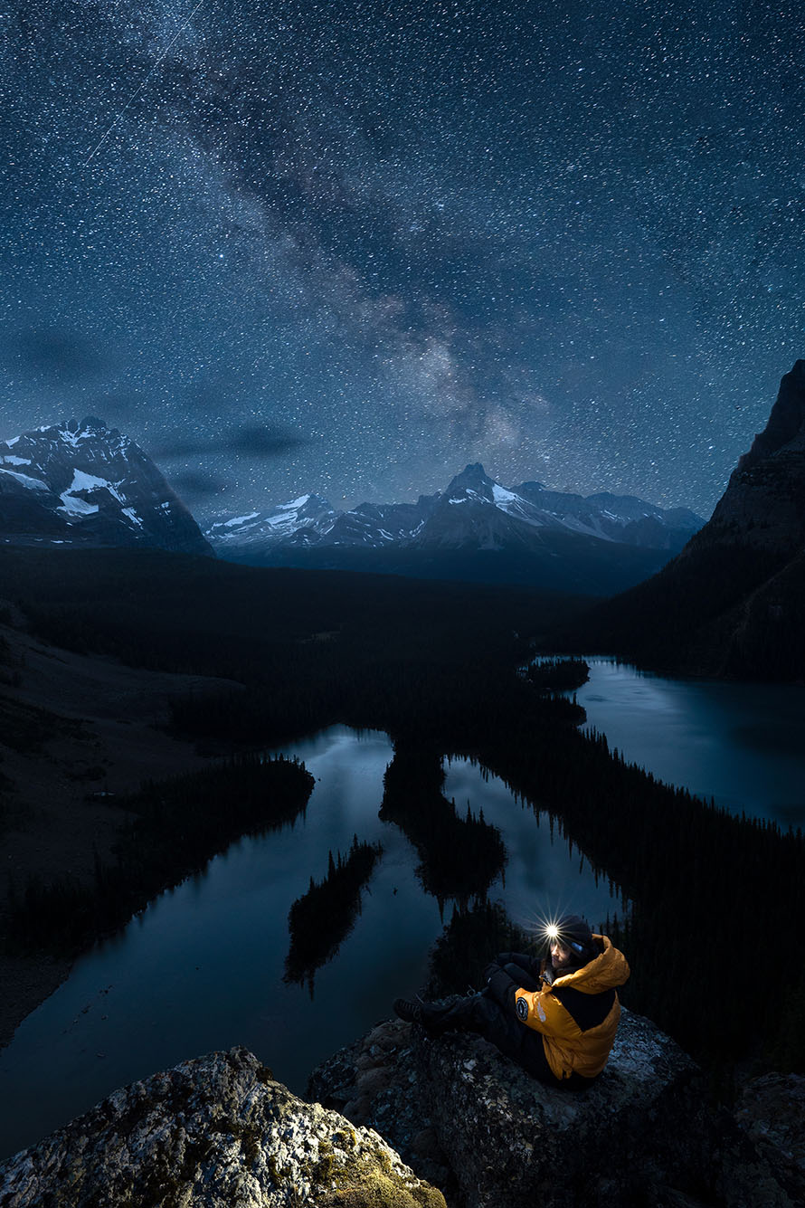Photo: Large, dramatic photo of a starry night sky over scenic mountains. A person can be seen in the foreground wearing a yellow puffer jacket and a headlamp that illuminates him and the area around him. Bodies of water can be seen around dark trees below the cliff the man sits on.