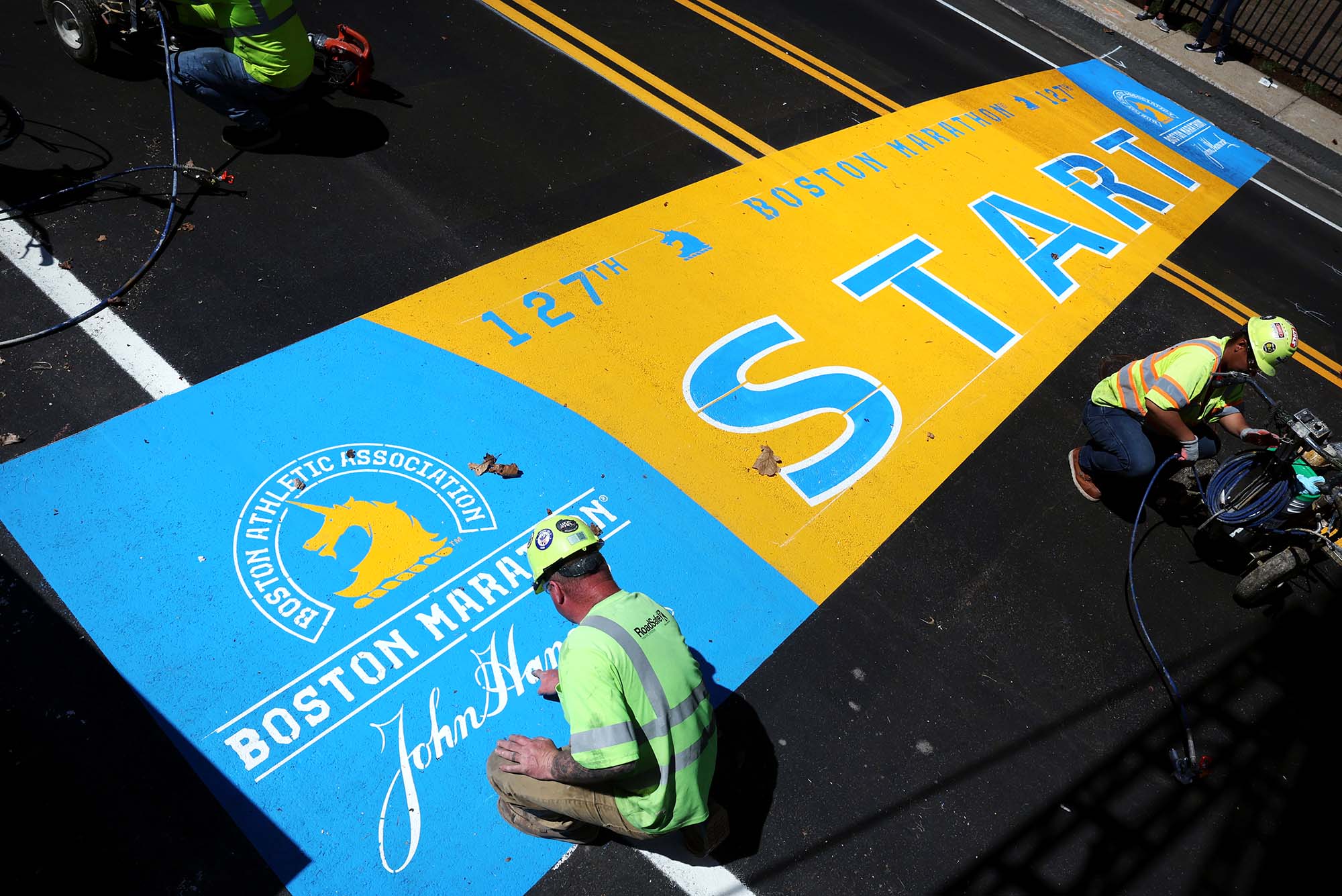 Photo: James Sawler, of RoadSafe Traffic Systems, paints the starting line for the Boston Marathon. A man wearing a white hard hat, neon shirt, and work gear paints on the light blue and yellow finish line on the road.