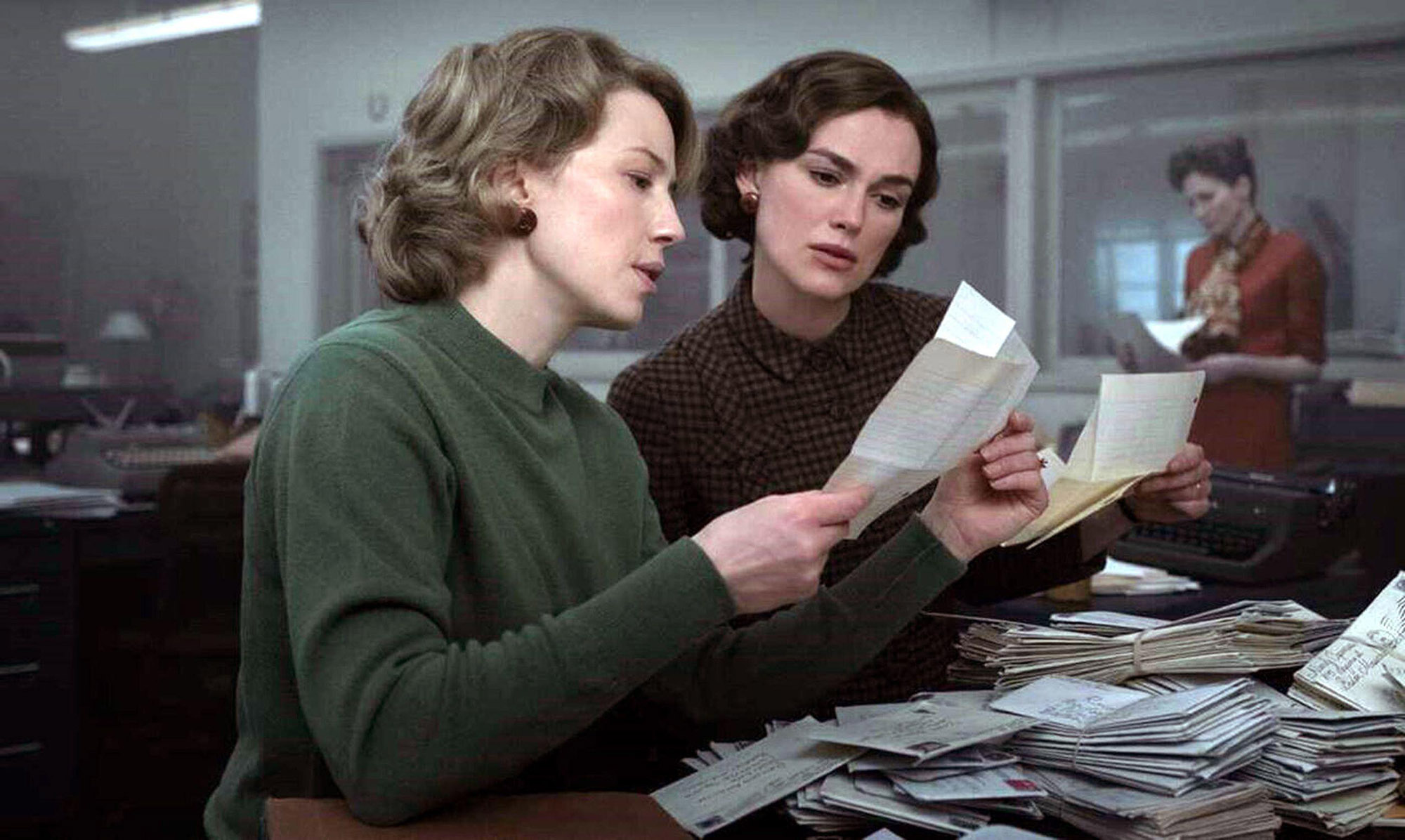 Screenshot from the Boston Strangler American historical crime drama film written and directed by Matt Ruskin. Keira Knightley as Loretta McLaughlin (right) looks over papers shown to her by Carrie coon as Jean Cole. They both sit in front of a desk piled with papers in an old-school newsroom.