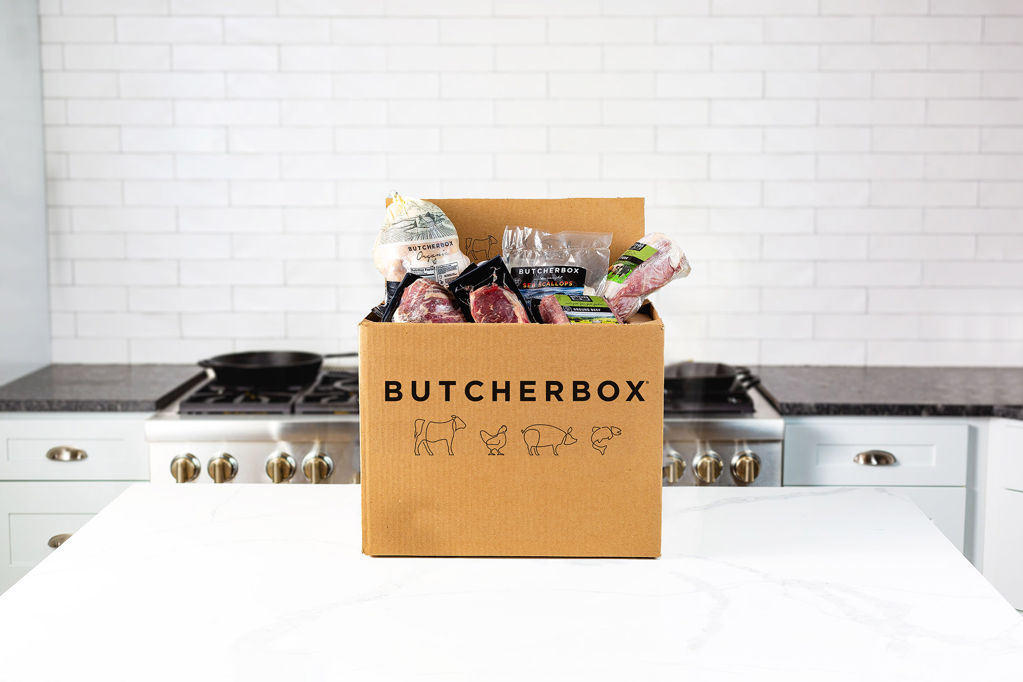 Photo: A brown box with the word "Butcherbox" and illustrations of a cow, chicken, pig, and fish are on the front of the box in black. The top of the box is overflowing with various meat products, like ground beef, steaks, and fish filets.