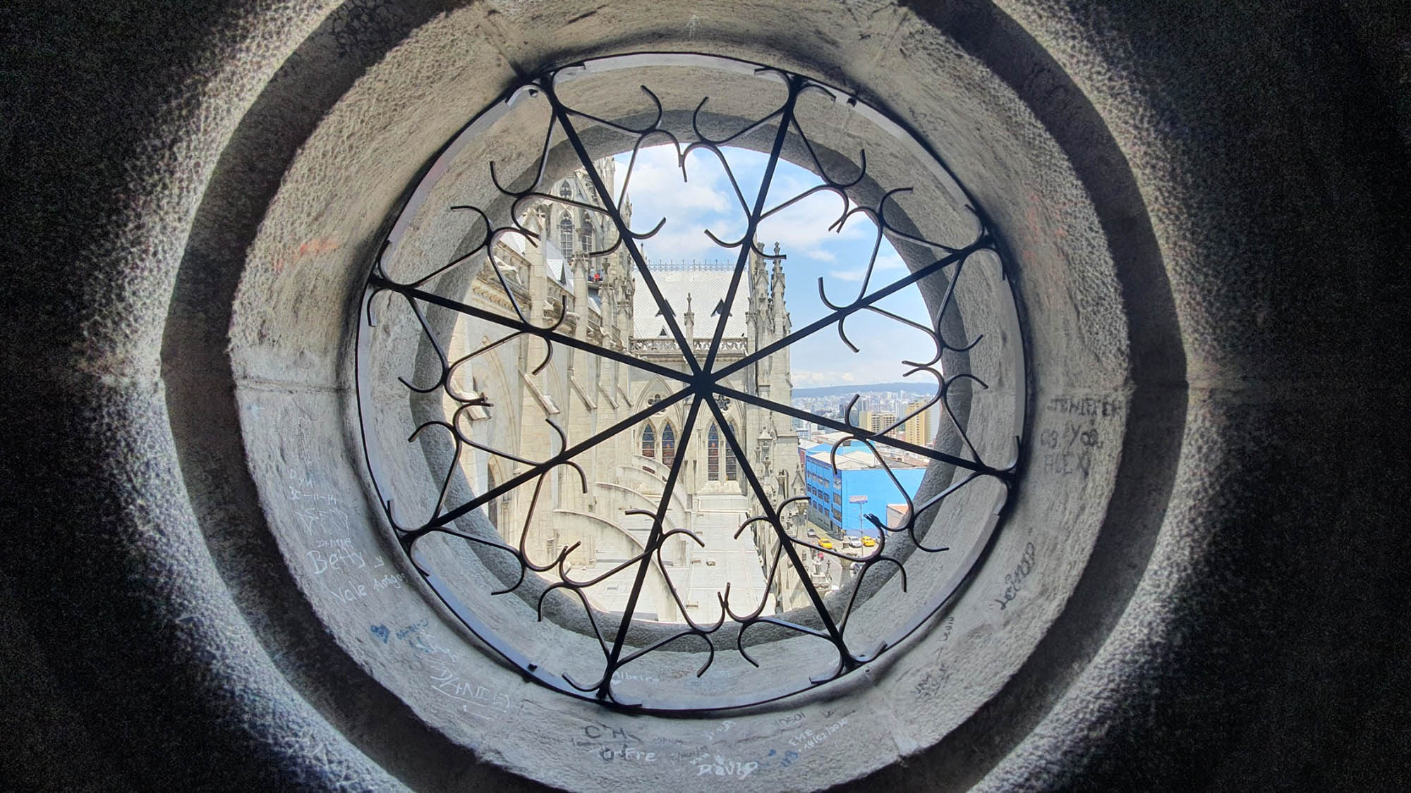 Photo: Shot taken from inside an observation window high up in The Basilica of the National Vow. A small circular window with an ornate gate covering is shown. Outside the window a a large basilica near a large body of water can be seen.