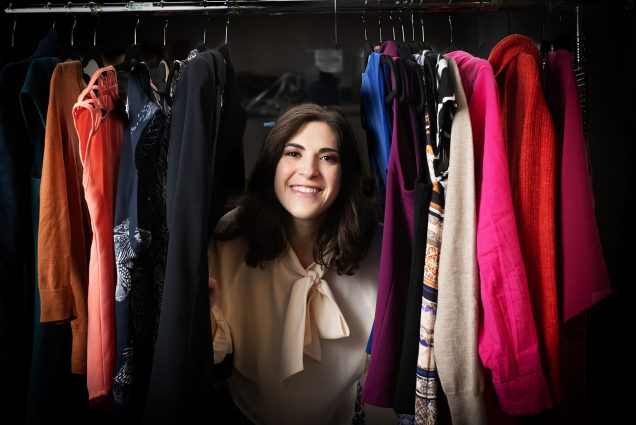Photo: Isabel Novick, a white woman with wavy brown hair and wearing a cream long sleeved blouse smiles as she peers out from the other side of a clothes rack. She pushes aside a set of clothes and poses from in between them.
