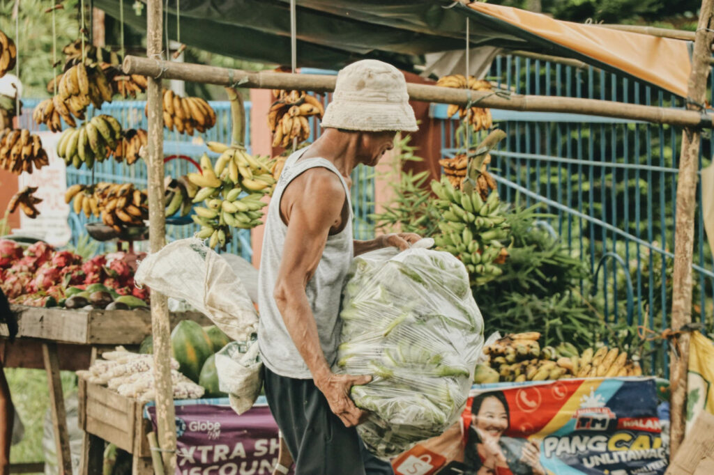 Photo:  a street vendor wearing a beige bucket hat, light grey tan top, and black sweatpants carries a large plastic bag filled with green bananas down a busy street. A fruit stall is seen behind him, displaying hanging green and yellow bananas as well as dragonfruit.
