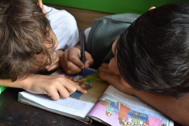 Photo: Two children, one white with light brown hair and the other a dark tan with black hair, look over a book together. They each lay their head down on the table and book. The white child points to something on the page as the dark tan child uses a blue crayon to mark something on the book.