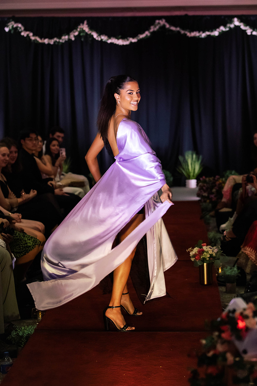 Photo: Reeya Mathur (QST’25) styled and wearing designs by Anabelle Pinkston (QST’23) at the BU’s Fashion & Retail Association annual fashion show. A young, tan woman with a slicked back, long ponytail and wearing a lavender drapey dress smiles as she twirls and looks back at the camera. She walks on a red runway with a spotlight as audience members sit on either side and watch in the darkened room.